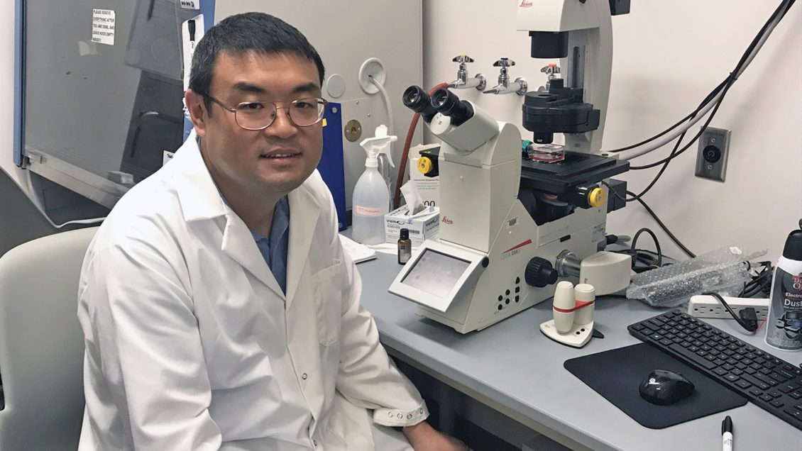 Zhicheng Dou sitting at microscope in his lab