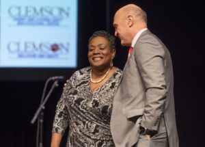 President Jim Clements stands in front of a smiling Deon Legette on stage in front of a video scree the Clemson University logo