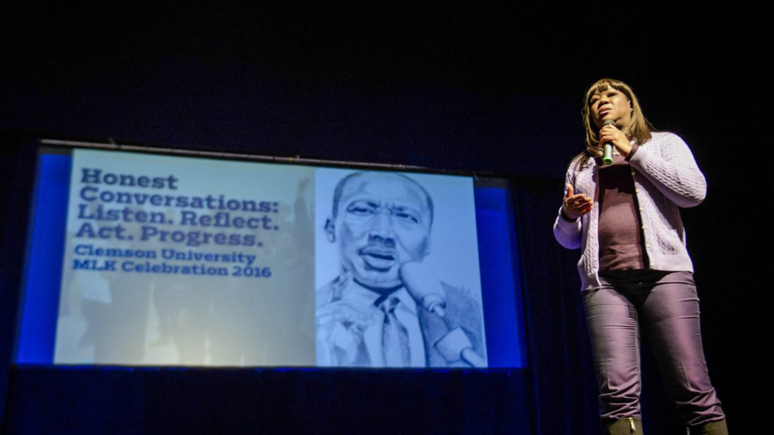 A woman stands on stage speaking into a microphone with a screen showing a graphic of the face of Dr. Martin Luther King Jr.