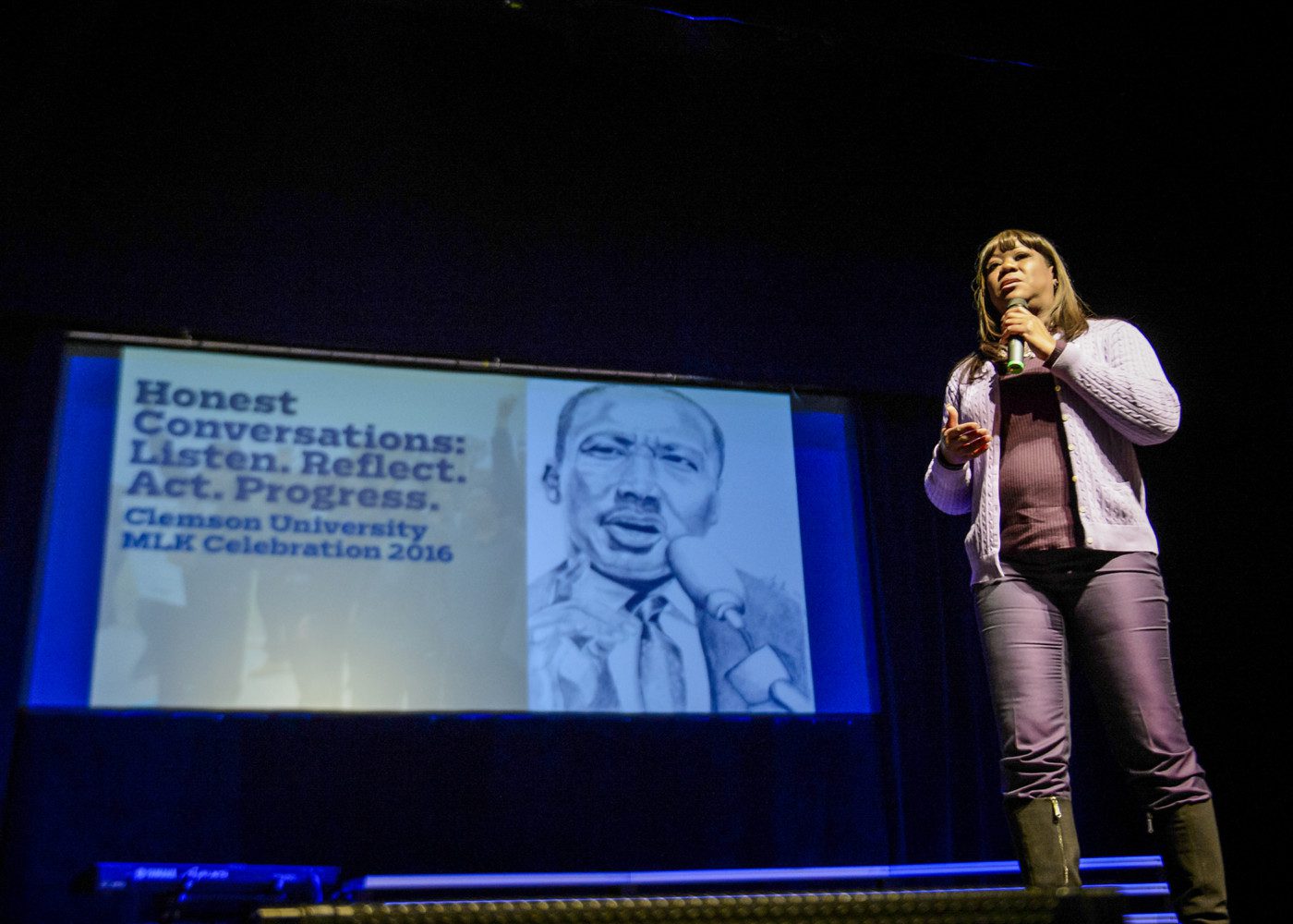 A woman stands on stage speaking into a microphone with a screen showing a graphic of the face of Dr. Martin Luther King Jr.