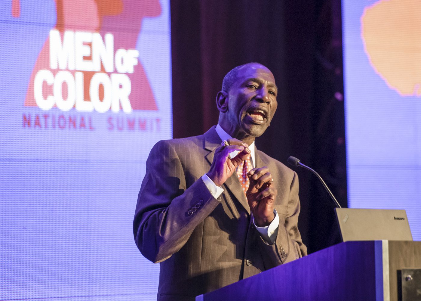 A dapper gentleman in nice suit gestures and speaks in front of a screen that says "Men of Color National Summit"