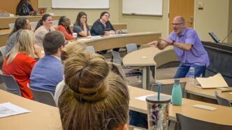 A teacher gestures stands at the front of a classroom and gestures to his students