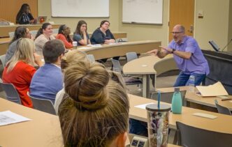 A teacher gestures stands at the front of a classroom and gestures to his students