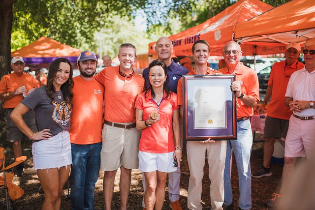 Seven people, Candi Glenn, her son, his girlfriend, and her husband are standing with President Jim Clements, Mike Dowling, and Will Brasington are at a Clemson University tailgating party. Candi is receiving her framed honorary alumni award.