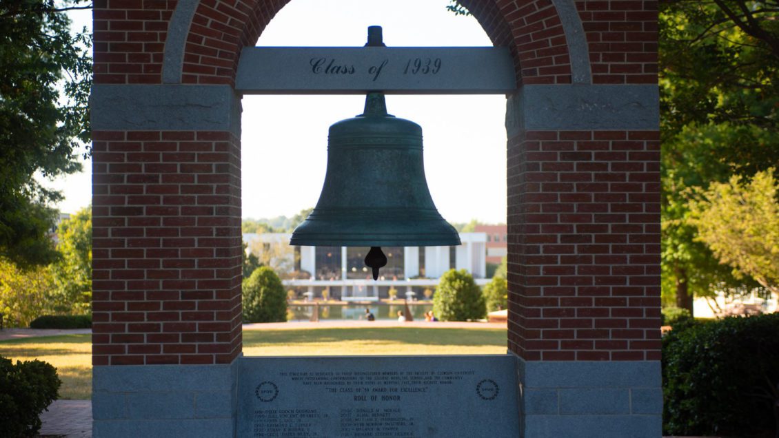 A big bell hangs in the middle of a plaza overlooking a library building in the distance