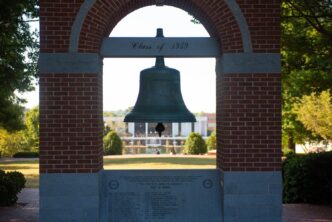 A big bell hangs in the middle of a plaza overlooking a library building in the distance