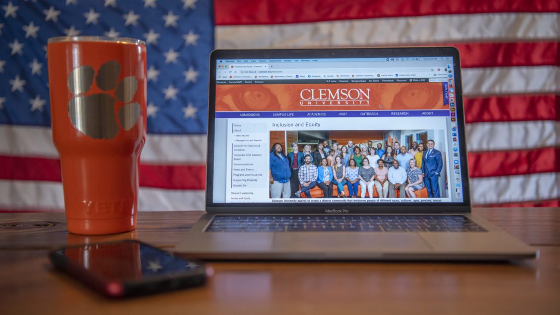 A laptop with the Division of Inclusion and Equity home page on its screen sits on a wooden table with a Clemson cup next to it and an American flag in the background.