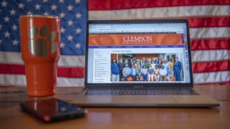 A laptop with the Division of Inclusion and Equity home page on its screen sits on a wooden table with a Clemson cup next to it and an American flag in the background.