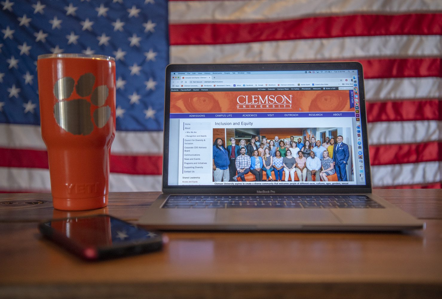 A laptop with the Division of Inclusion and Equity home page on its screen sits on a wooden table with a Clemson cup next to it and an American flag in the background.