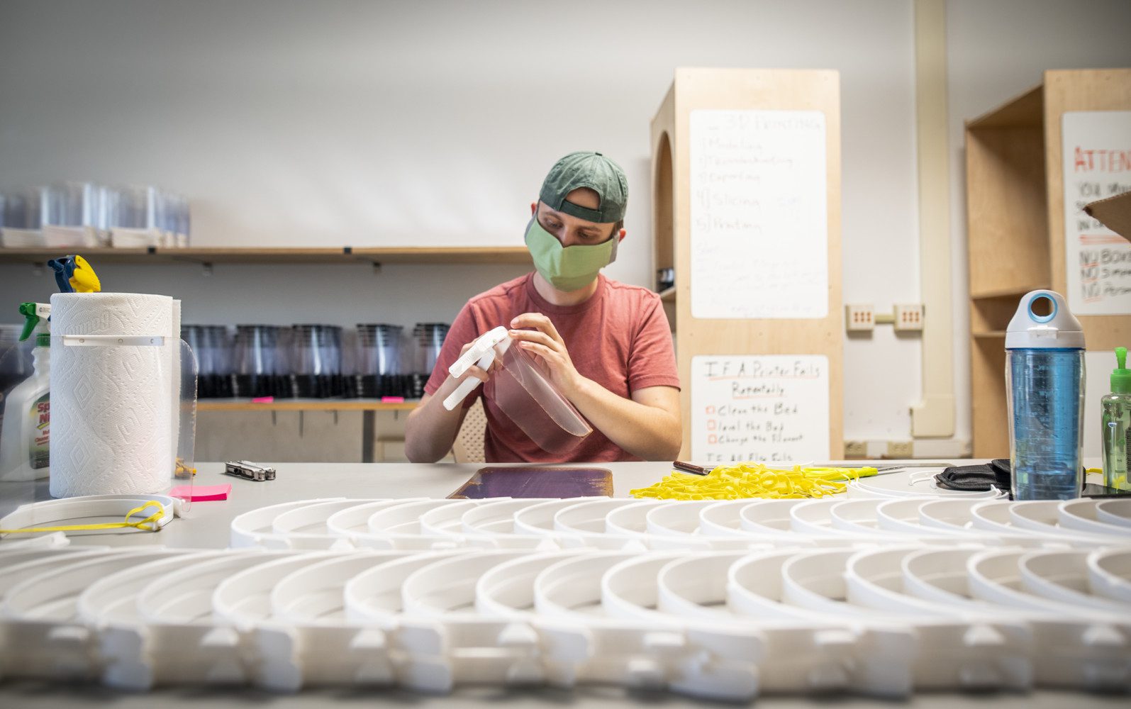 A student wearing a cloth face mask works at a table covered with rows of face shield parts