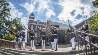 Sun beams shine through a formation of soldiers who are raising their rifles and firing in front of Tillman Hall.