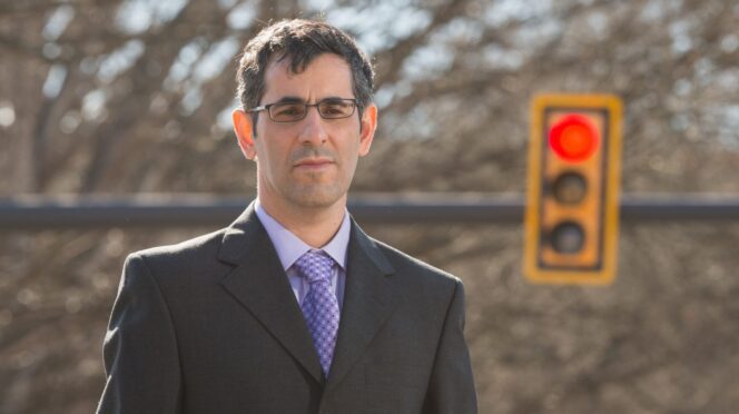 A man in a suit stands outside with a stop light behind him.