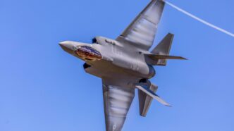A fighter jet streaks through the blue sky, tipped sideways toward the camera