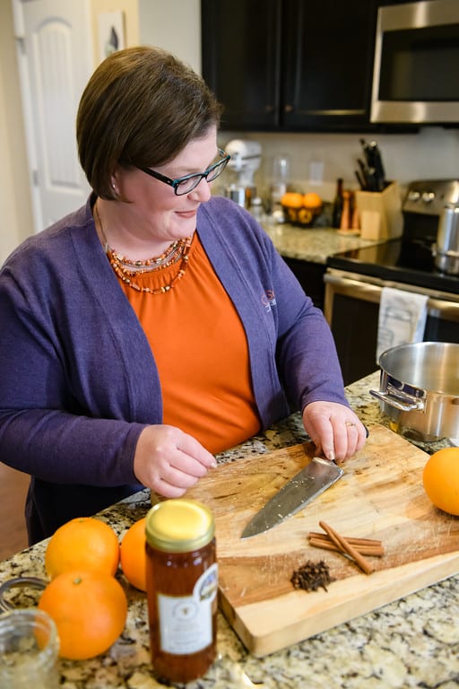 Woman (Kim Baker) wearing glasses and a blouse and cardigan with a two-strand beaded necklace is smiling and standing in a kitchen behind a counter, holding knife and crushing spices on wooden cutting board. Three oranges sit on the side of the cutting board, and one orange is on top. Fruit, and its juice, can be a substitute for sugar.