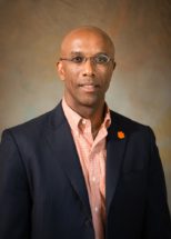 A smiling man wearing small framed glasses, sports jacket and button down shirt with a Clemson insignia tiger paw lapel pin
