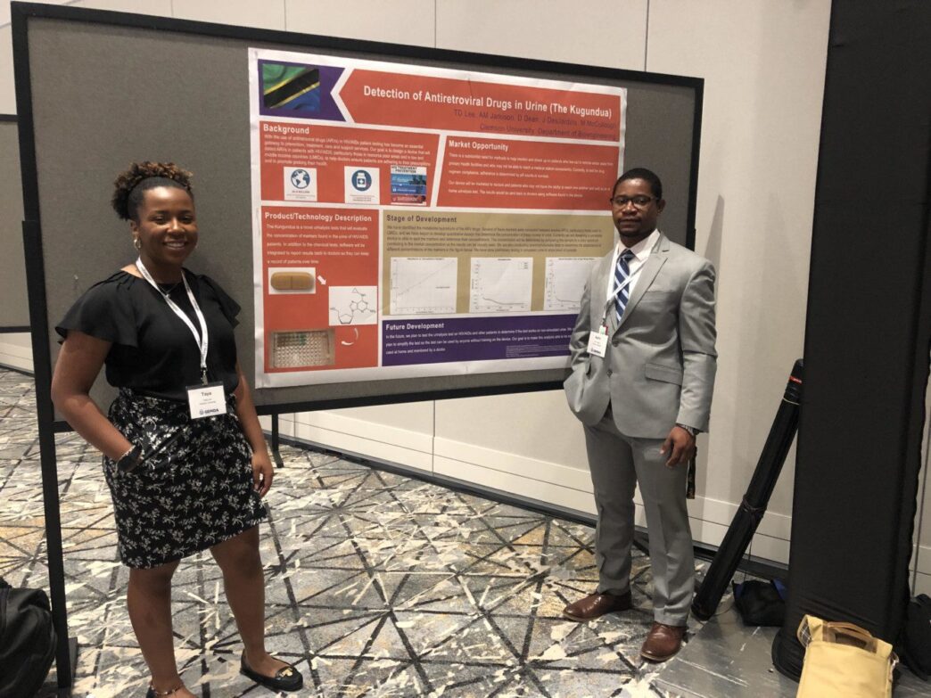 Two smiling students in business attire standing in front of a presentation board that displays information about their project