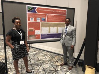 Two smiling students in business attire standing in front of a presentation board that displays information about their project