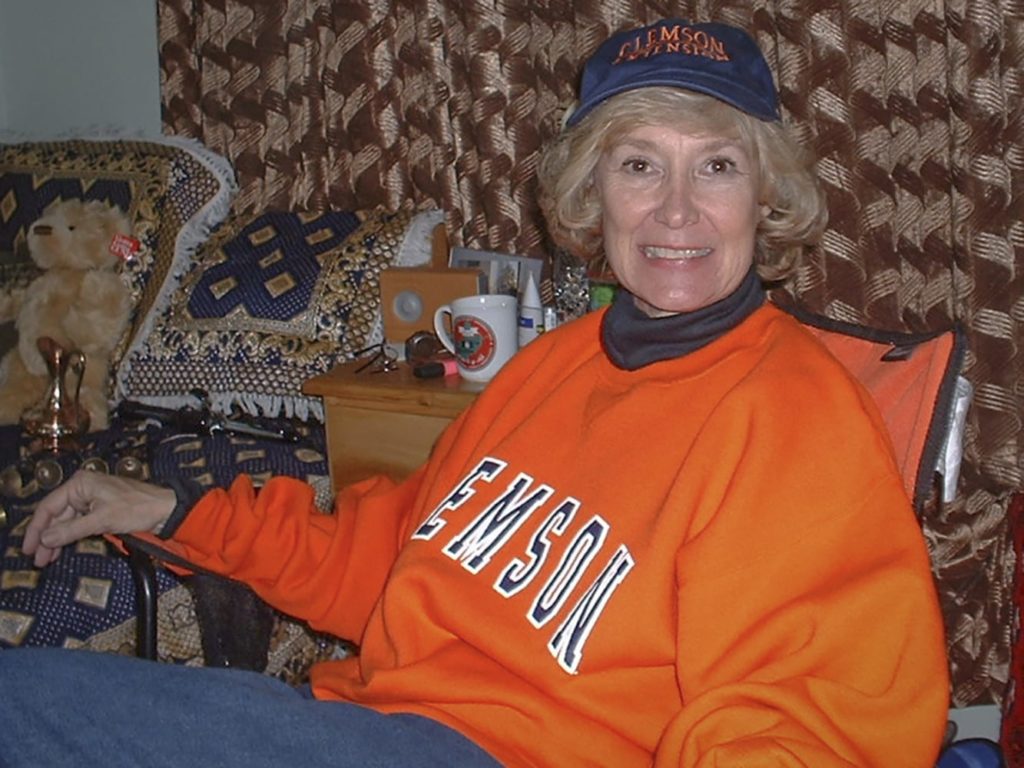 A woman wearing a Clemson orange sweat shirt and Clemson ball cap sits in a small room, in front of muted striped curtains