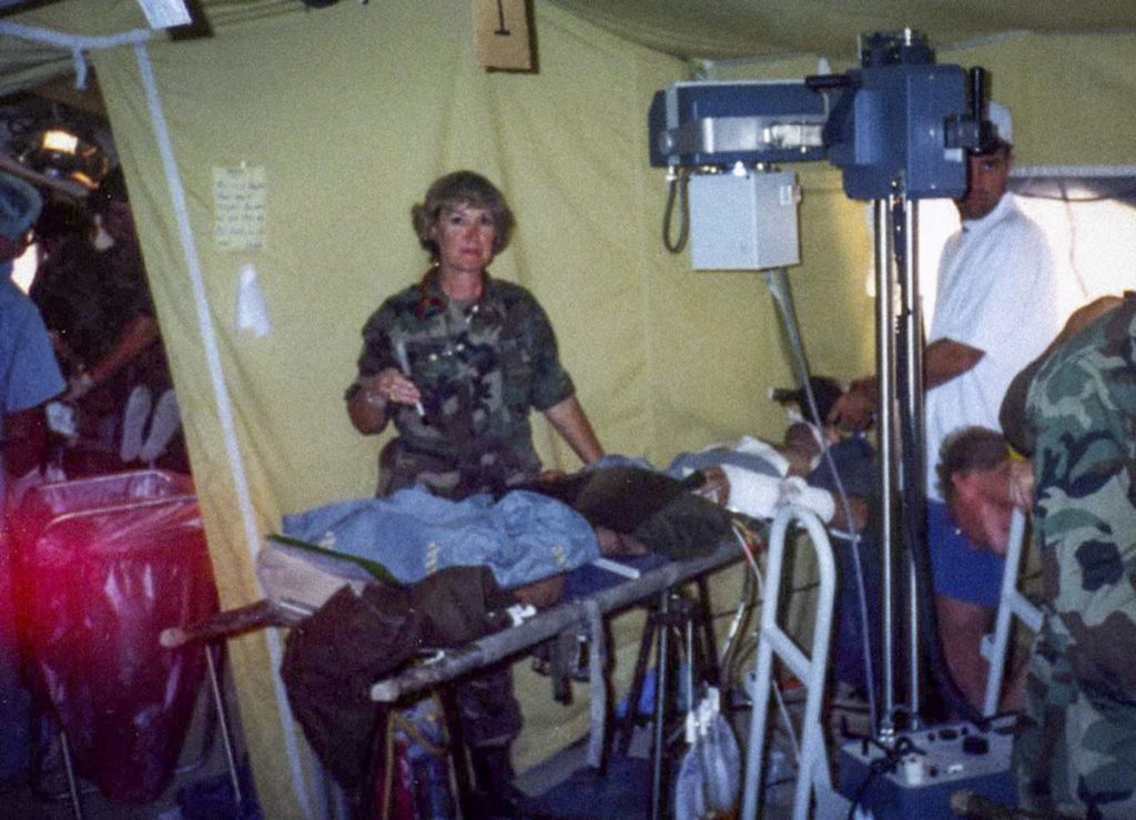 A woman in Army uniform standing next to a stretcher, in a tent, with medical equipment all around her