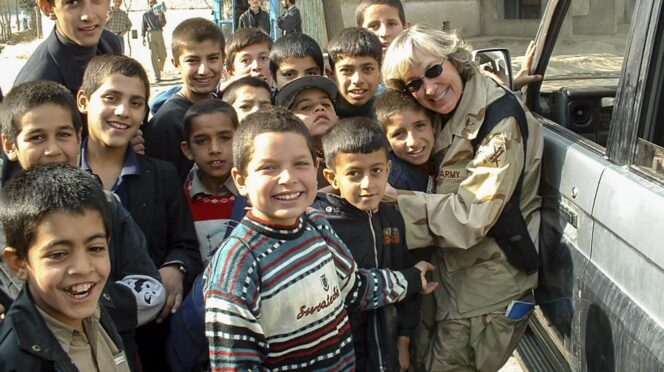 A female Army officer in uniform embraces a young child while a dozen other children crowd around her
