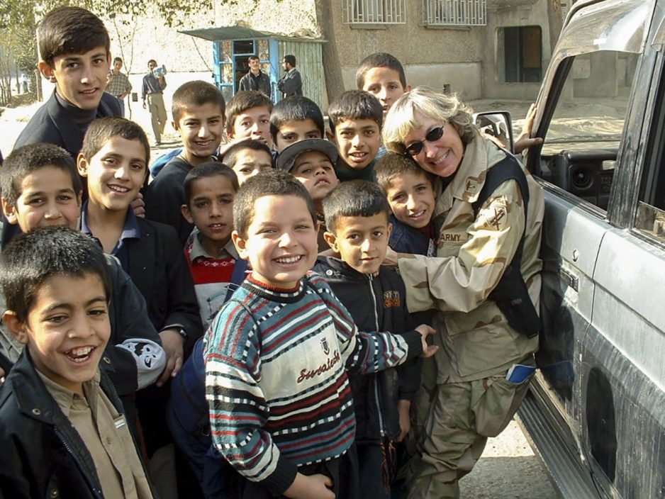 A female Army officer in uniform embraces a young child while a dozen other children crowd around her