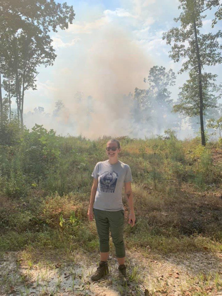 Crystal Smith standing in the woods with smoke behind her.