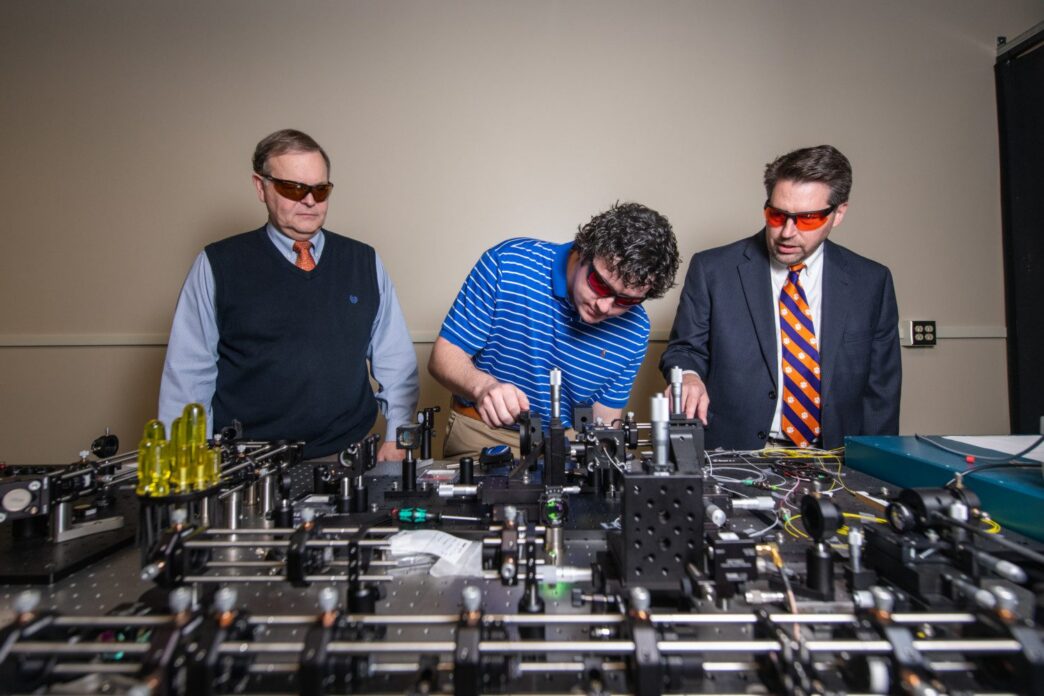 Three men in business attire looking at a project in an engineering lab.