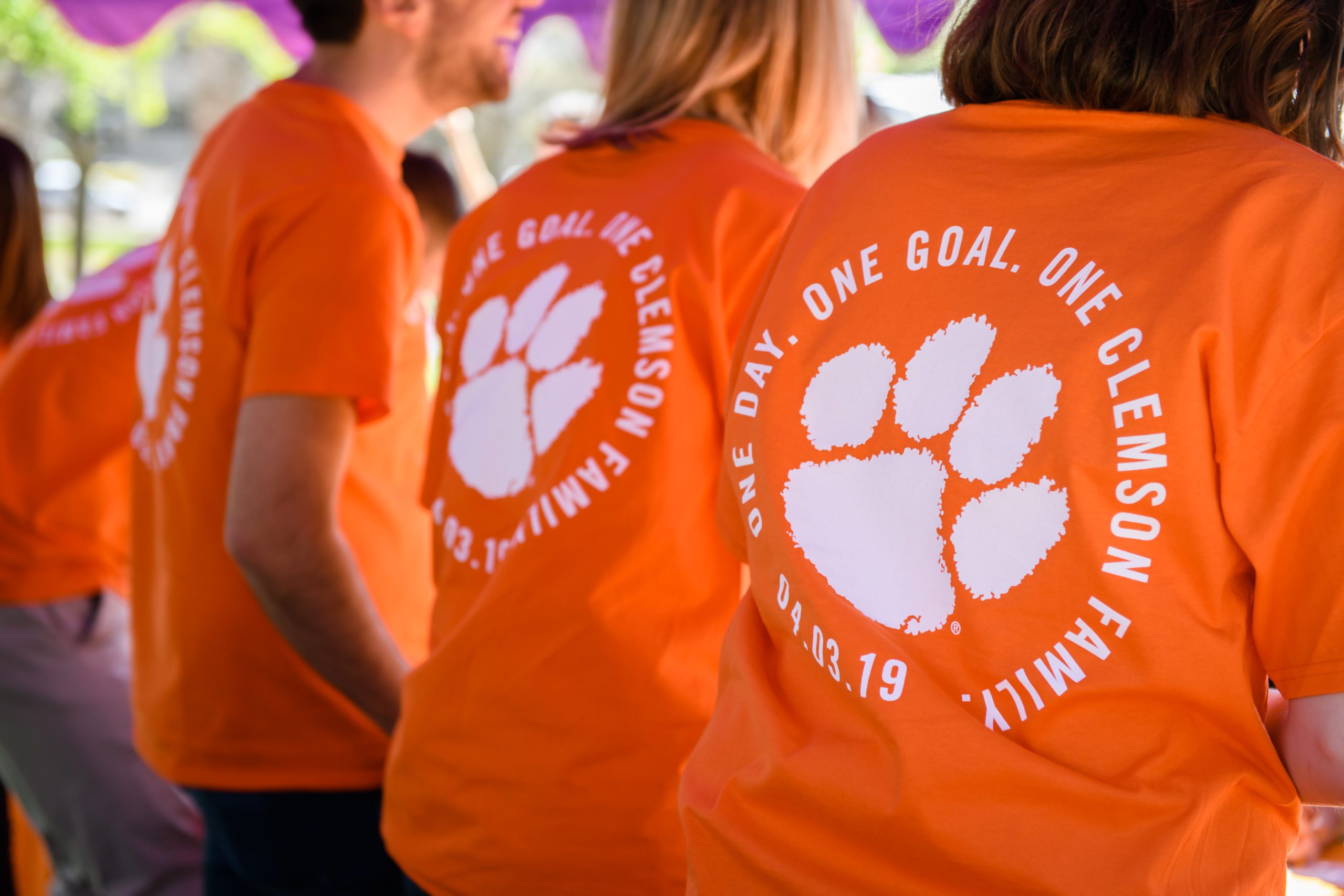 Showing the back of three individuals standing with One Goal. One Clemson on their shirt.