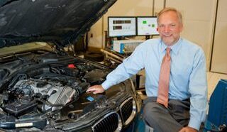 Zoran headshot seated in front of a car with the hod up so you can see the engine.