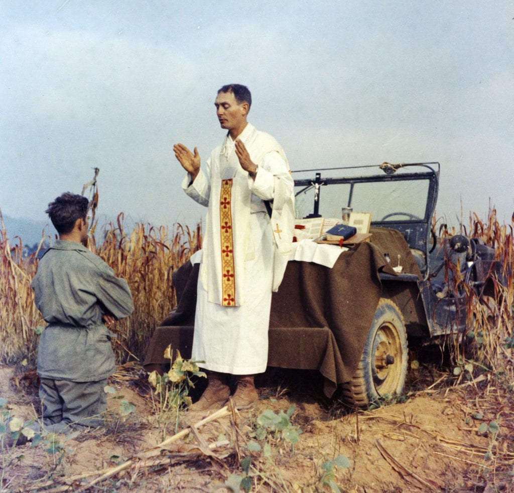 Photo of a chaplain in robes standing next to a jeep, a soldier kneeling in front of him