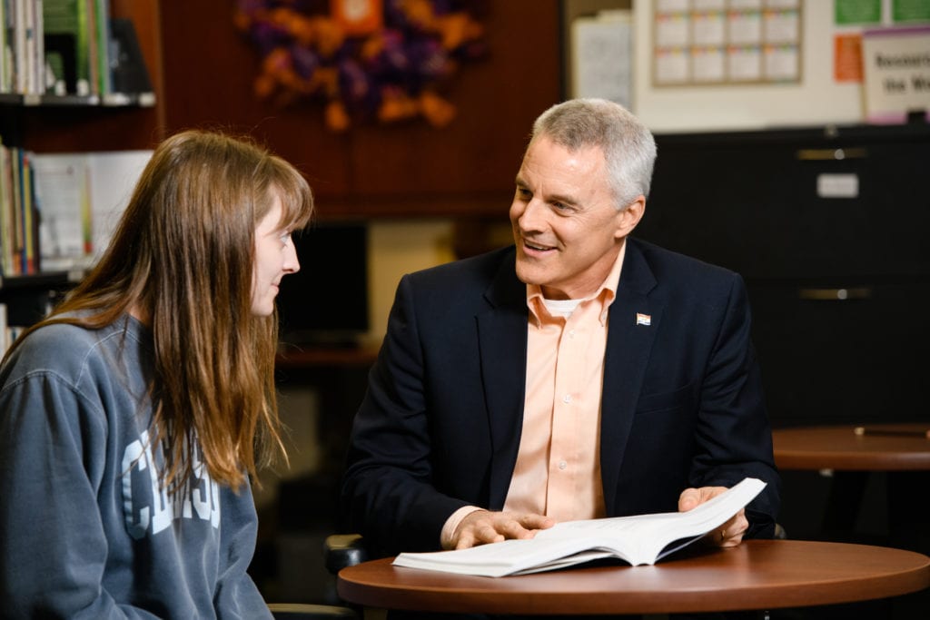 Man in black suit with orange shirt sitting at table looking at a book with a female student