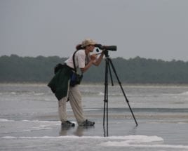 woman with a hat standing on a beach looking through a scope