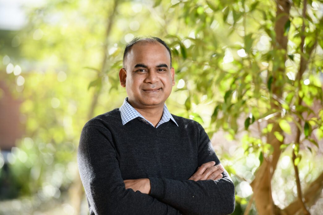 A man stands with his arms crossed in front of green trees.