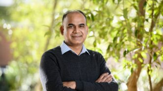 A man stands with his arms crossed in front of green trees.