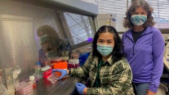 Female student with mask sitting at lab table with professor Lisa Bain standing near her