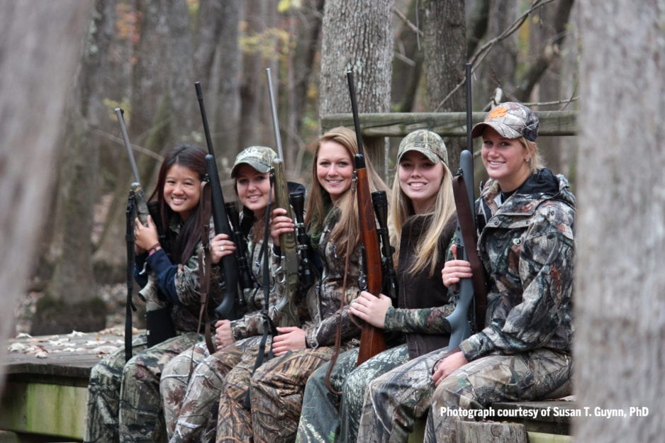 Five women smile as they sit in a line wearing camouflage and holding guns.