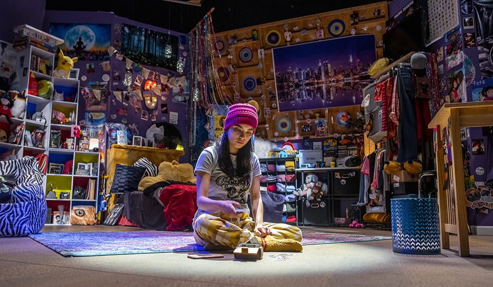 A female actor sits in a bright, cluttered scene for rehearsal. 