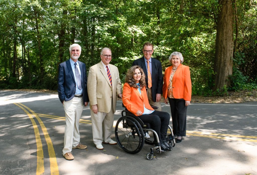 The Master of Transportation Safety Administration (MTSA) team, consisting of Bruce Rafert, Philip Pidgeon, Kim Alexander, Ralph Elliott and Terecia Wilson, are on a curved, tree-lined secondary road with intersecting yellow traffic lines. Alexander is sitting in her wheelchair with Rafert and Pidgeon standing by her right side and Elliot and Wilson on her left.