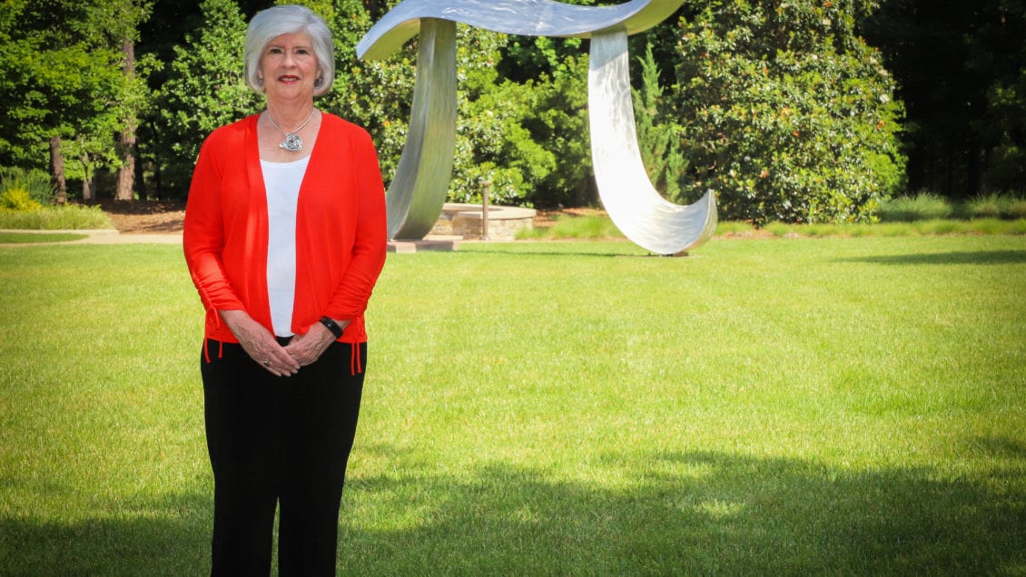 Emily Wallace, a Clemson graduate from the class of 1972, stands outdoors in front of a metal sculpture in the shape of a mathematical pi symbol. She is wearing business casual attire.