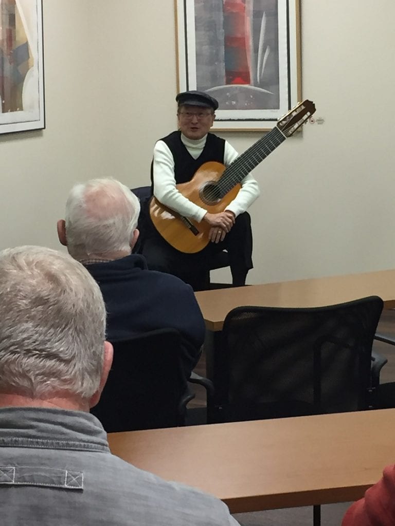 Yuji Kishimoto, professor emeritus of architecture, sits at the front of a room cradling a guitar in his lap with a small audience looking on. 