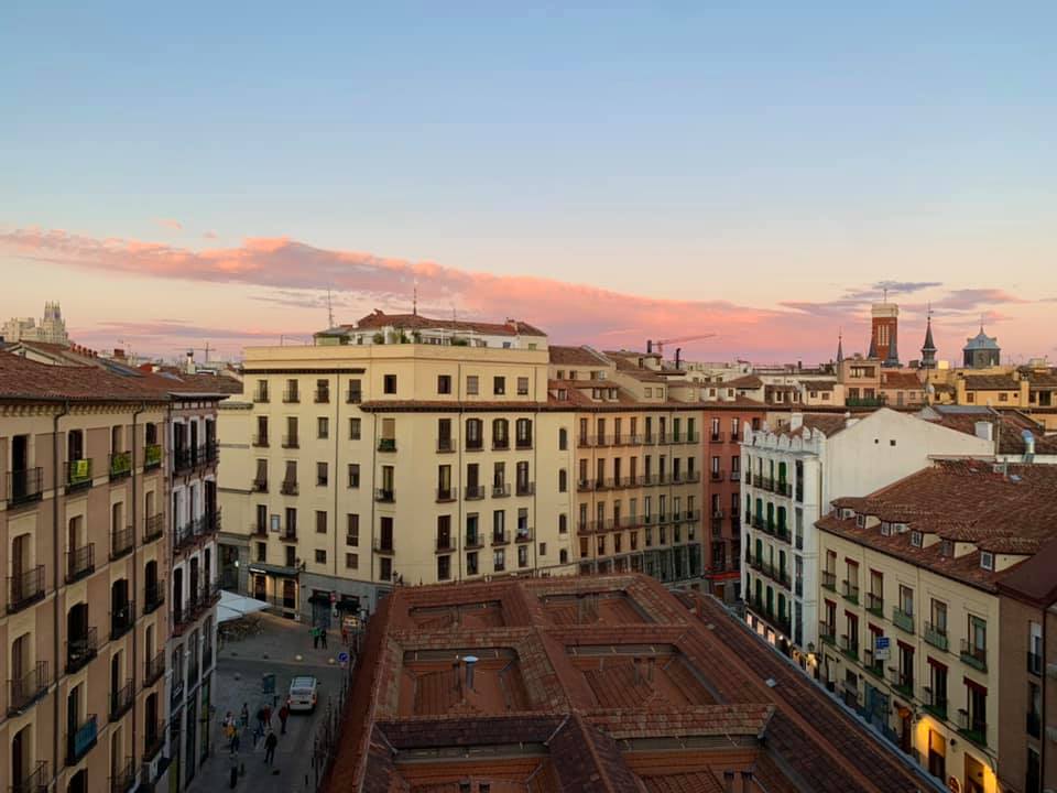An apartment view overlooking a residential area in Spain. There are clouds in the sky, above the horizon, and rooftops, steeples and hints at significant cultural and architectural landmarks in the distance.