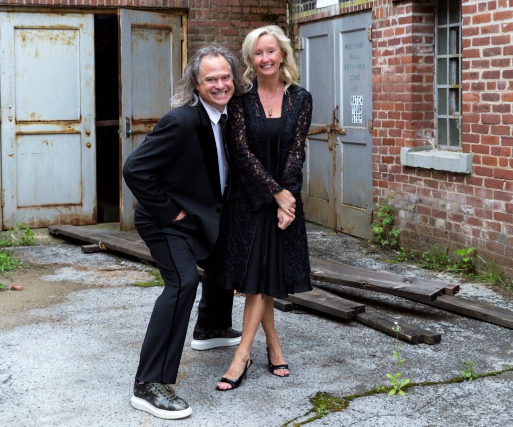 Man in his mid-fifties with with longer hair, wearing a tux and sneakers, is standing in a mid-crouch with his right hand in his front pocket and left arm around a woman in a cocktail dress, standing with her left leg crossed in front of her right. They are outside an old, neglected industrial brick warehouse with rusted steel doors. The both have happy, friendly smiles, and his smile looks a little mischievous. They are Clemson University alumni, husband Brook and wife Pam Smith of Kentucky, philanthropic supporters of entrepreneurial ventures.