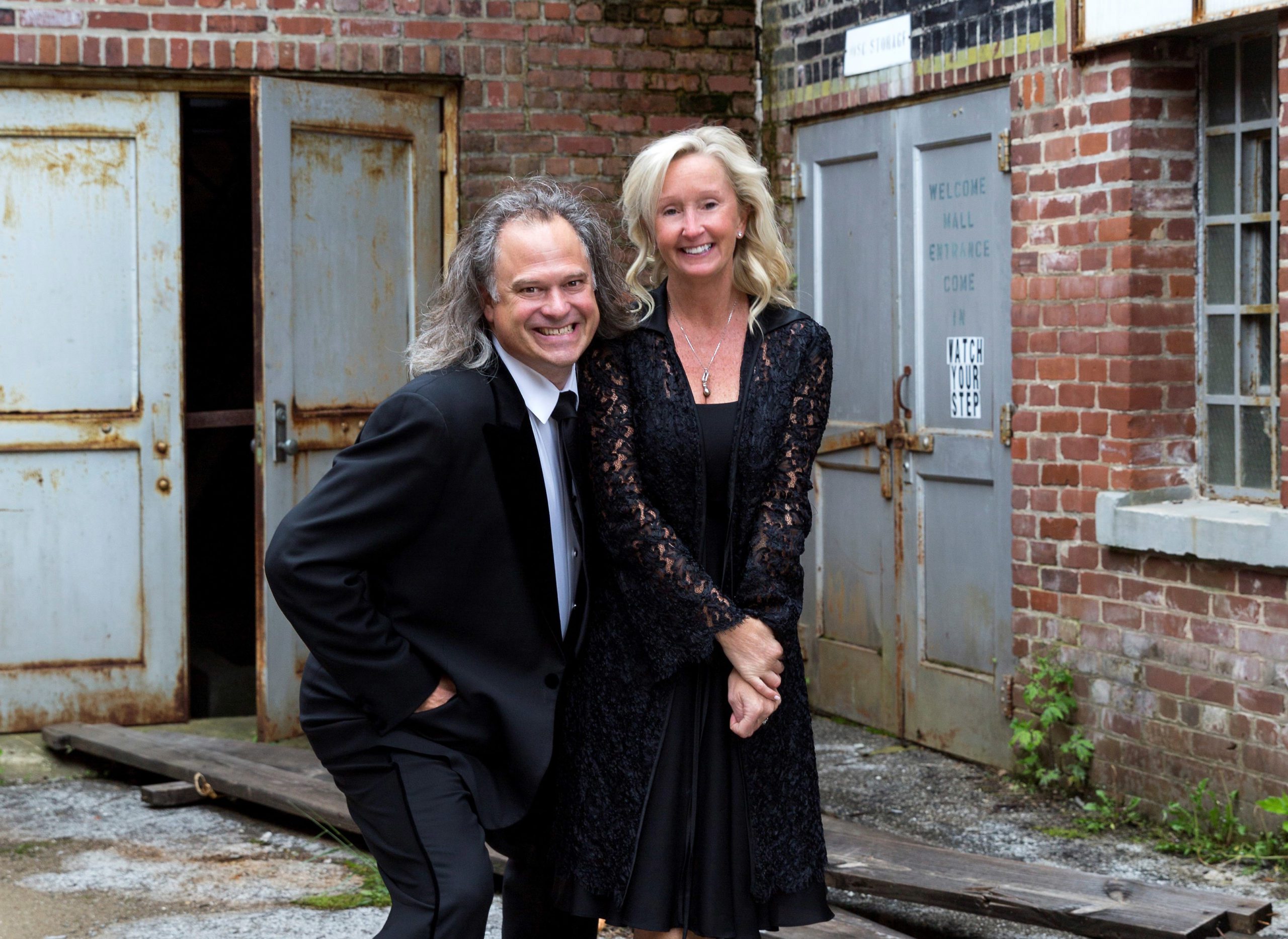 Man in his mid-fifties with longer hair, wearing a tux and sneakers, is standing in a mid-crouch with his right hand in his front pocket and left arm around a woman in a cocktail dress, standing with her left leg crossed in front of her right. They are outside an old, neglected industrial brick warehouse with rusted steel doors. The both have happy, friendly smiles, and his smile looks a little mischievous. They are Clemson University alumni, husband Brook and wife Pam Smith of Kentucky.