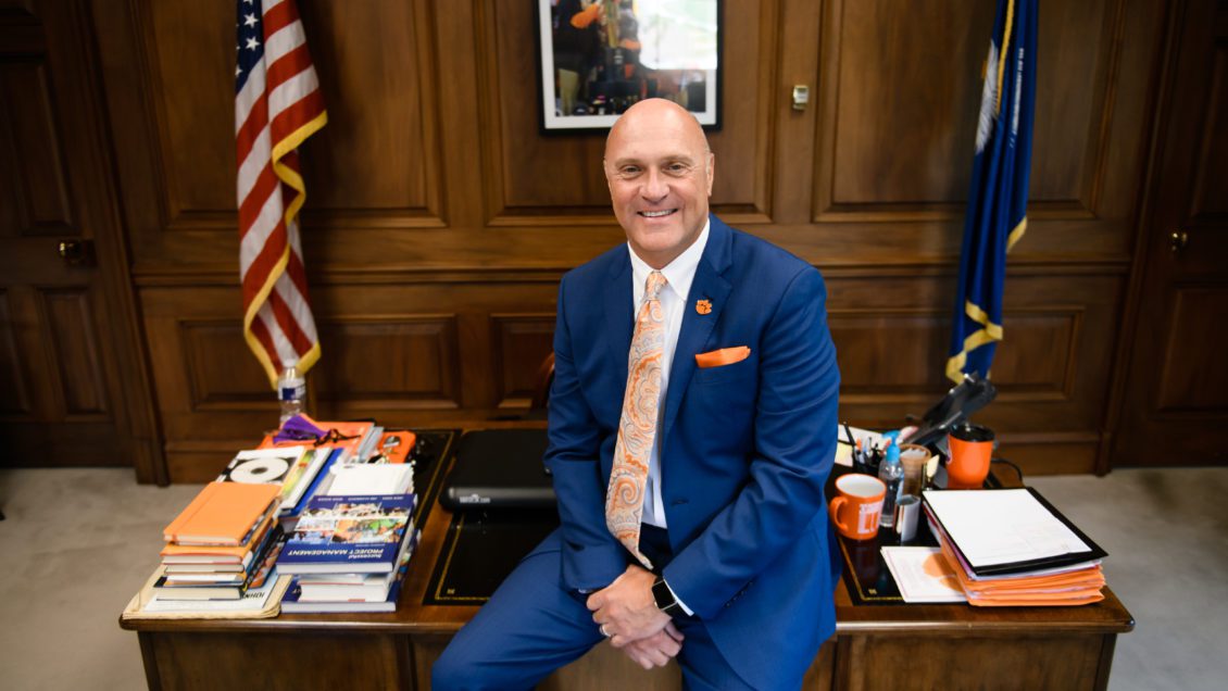 Clemson President Jim Clements is shown in a blue suit leaning against his desk.