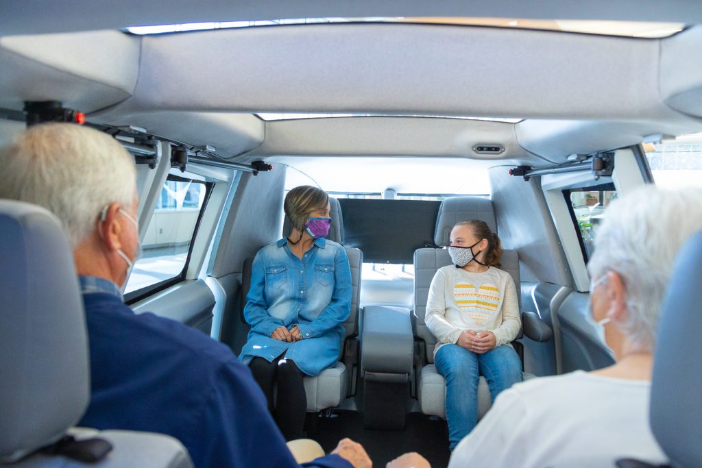 Four people sit in the light interior of a car. The two rows of seats face each other with legroom in the center. 