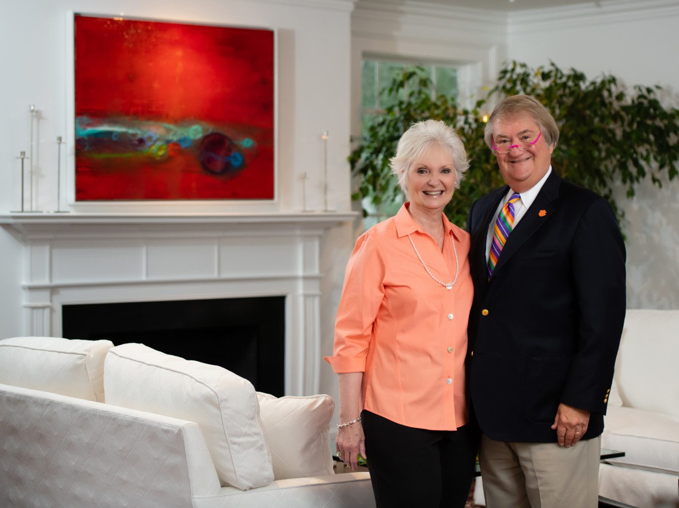 Image of Ben and Becca at home in Greenville. Becca in an orange shirt, Ben in a suit, standing in front of fireplace with large art piece on the wall.