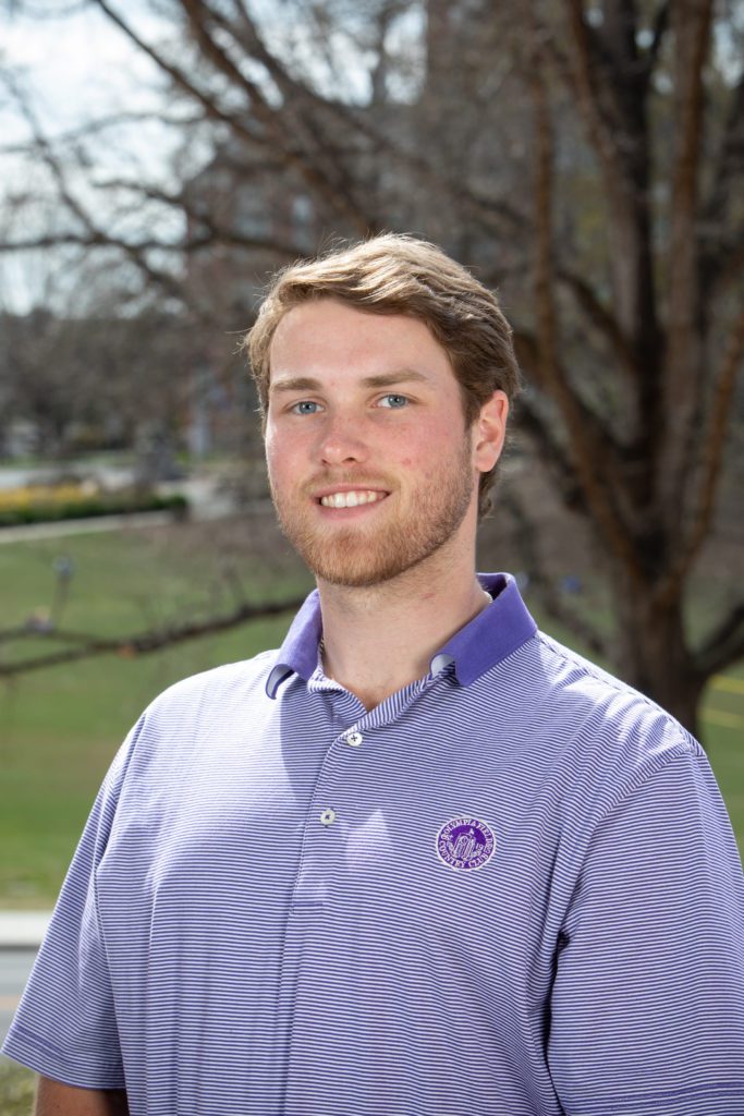 Student Caleb Hannon, a Powers Scholar, resulting from the philanthropic gift of Billy and Ann Powers, is standing outside. There is a tree and field behind him, along with a building in the far distance.