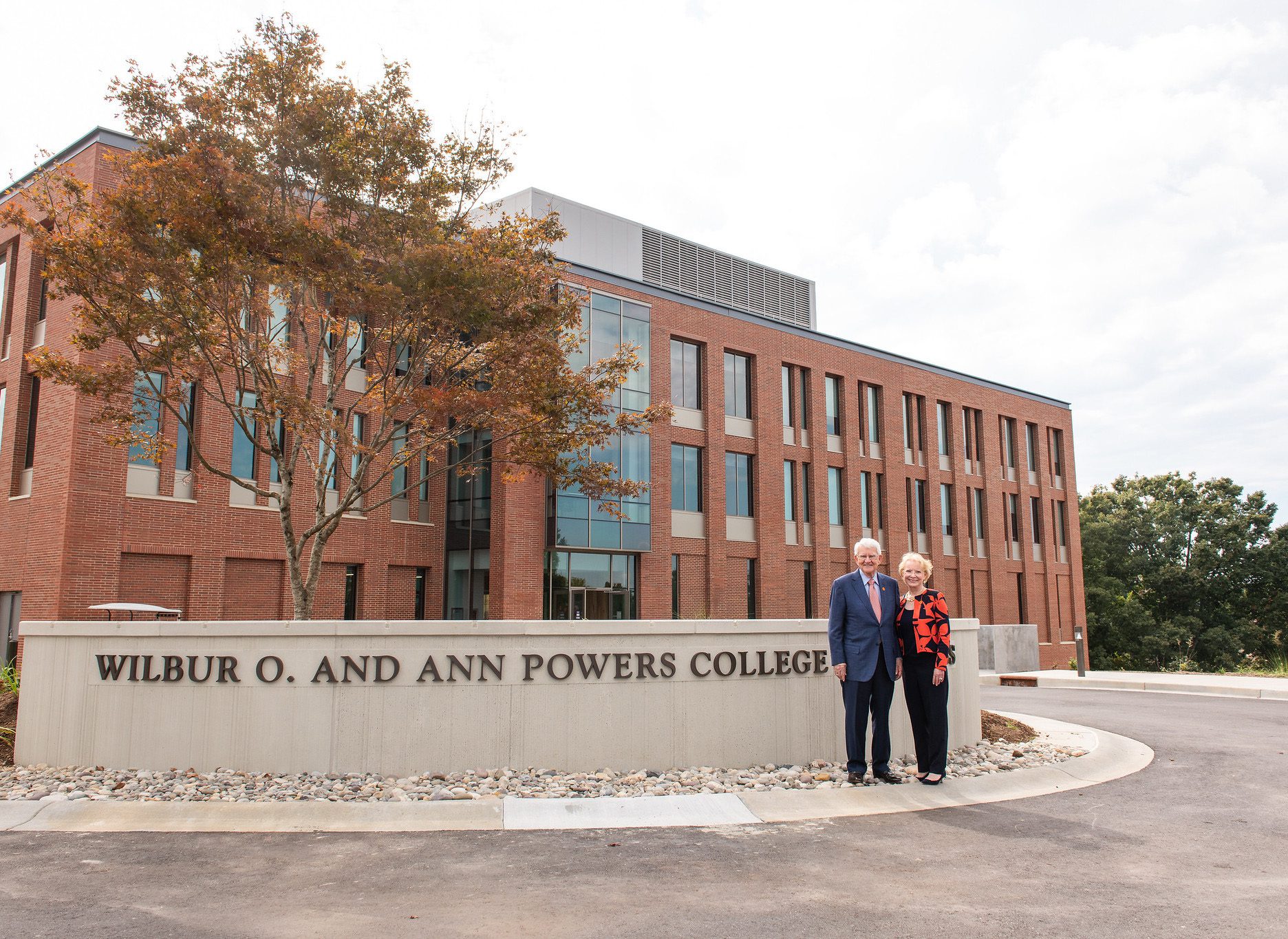 An older couple, philanthropists Billy and Ann Powers, are standing in front of a long, concrete sign that stand about four feet from the ground and is slightly curved towards a new brick and glass building. The building is a part of the newly built business school, and the sign reads Wilbur O. and Ann Powers College of Business, named after the philanthropic couple standing in front of it. The day is slightly windy and a little overcast. There is a rock garden in front of the sign that leads to a curb. Behind the sign, in front of the building, is a small garden with a tree as its center. The couple gave Clemson its largest philanthropic gift to date.