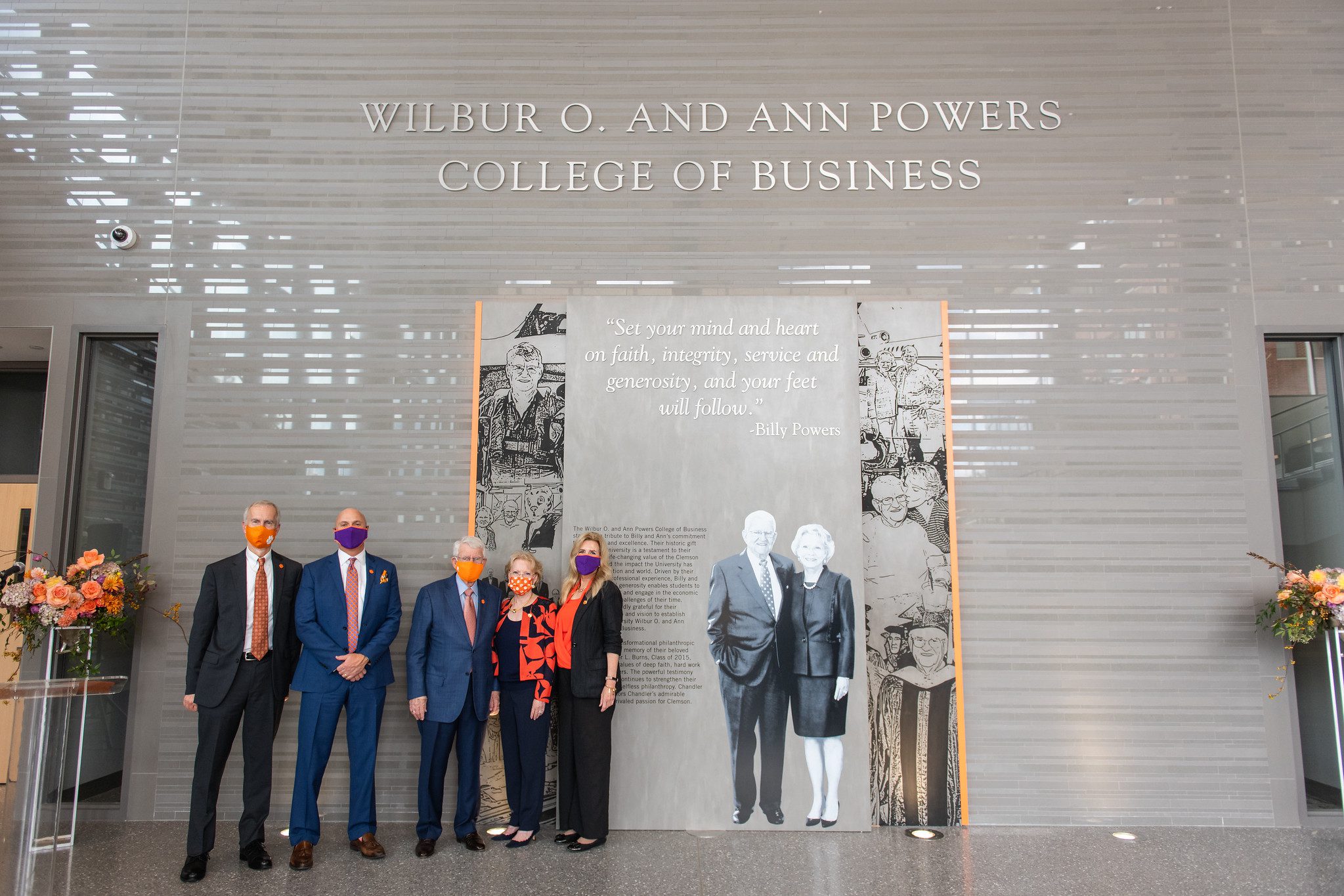 A group, including philanthropists Billy and Ann Powers, along with Dean York, is standing in front of a wall, inside a building, that features a quote and sketches of Billy and Ann Powers on its wall. Towering above the graphics is the name of the college, the Wilbur O. and Ann Powers College of Business, named to honor the philanthropic gift.