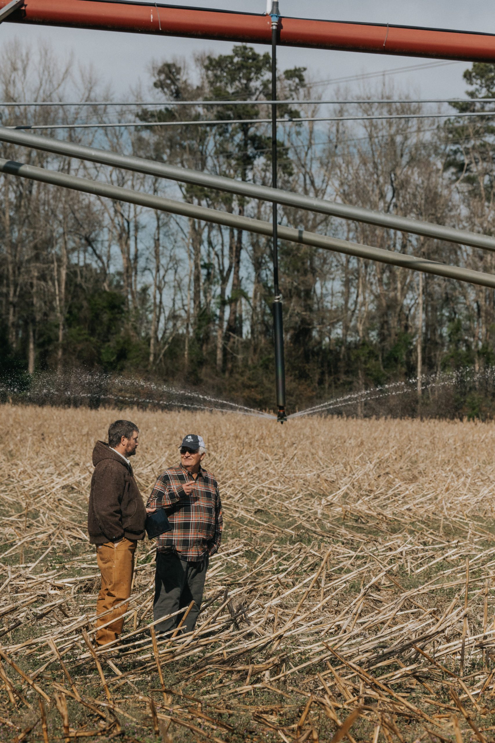 Clemson precision agriculture engineer Kendall Kirk and Bamberg County farmer Richard Rentz stand in a field as an irrigation sprayer works in the background. 